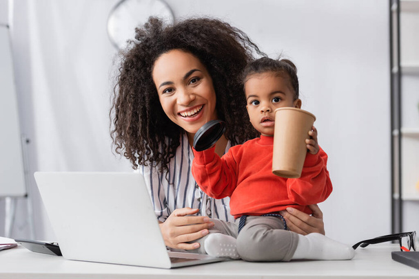 toddler african american child sitting on desk and holding paper cup near cheerful mother  - Photo, Image