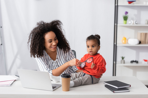 happy african american mother looking at daughter holding glasses while sitting on desk - Фото, изображение