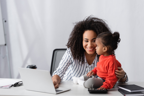 sonriente africano americano madre mirando a niño hija sentado cerca de portátil en escritorio - Foto, Imagen