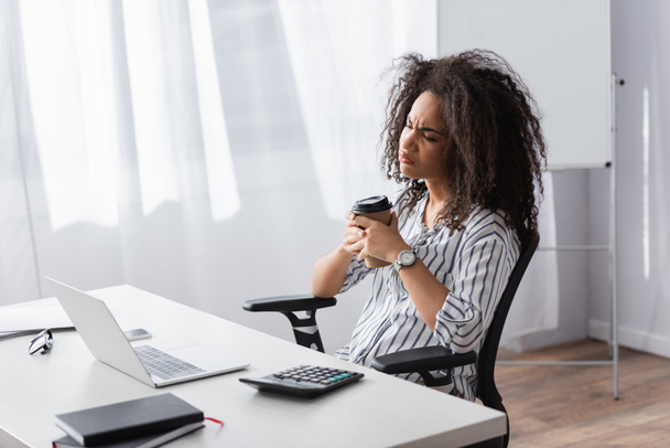 disgustado afroamericano freelancer sosteniendo taza de papel cerca de la computadora portátil en el escritorio  - Foto, imagen