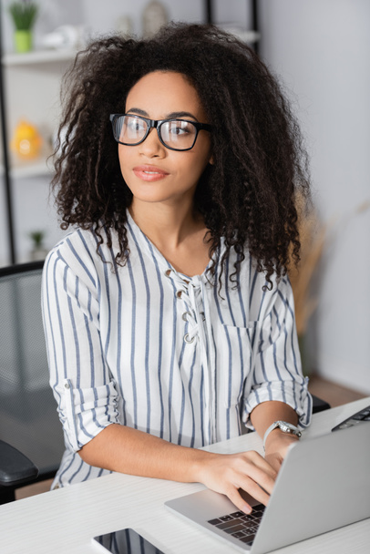 african american freelancer in glasses looking away and using laptop at home - Photo, Image