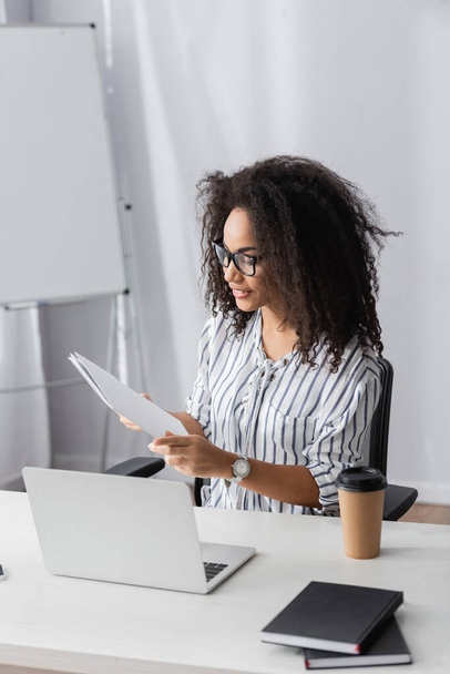 happy african american freelancer in glasses holding folder near laptop and paper cup on desk - Foto, Bild