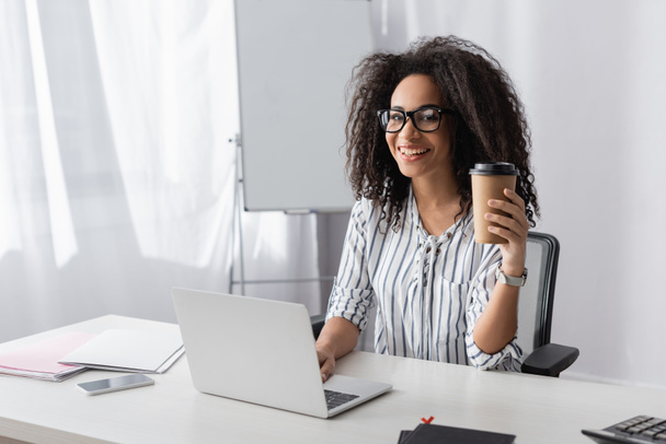 heureux afro-américain pigiste dans des lunettes tenant tasse en papier avec café pour aller près de l'ordinateur portable à la maison  - Photo, image