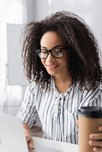 mulher americana africana feliz em óculos perto de xícara de papel com café para ir em primeiro plano borrado - Foto, Imagem