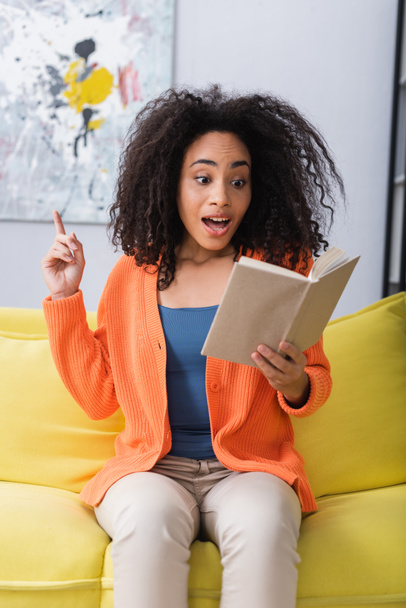 amazed african american woman having idea while reading book and sitting on couch - Photo, image