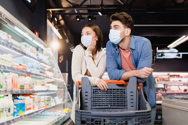 Paar mit Arztmasken betrachtet Lebensmittel in der Nähe von Einkaufswagen im Supermarkt  - Foto, Bild
