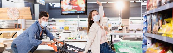 Man in medical mask rolling shopping trolley with girlfriend in supermarket on blurred foreground, banner  - 写真・画像