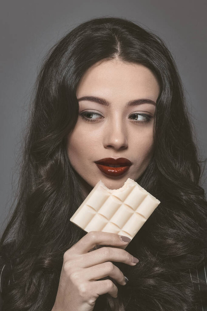 Sensual beautiful brunette young woman bite white bubbly chocolate wearing black jacket with beautiful long dark hair isolated on grey background. Young female model with professional make-up - 写真・画像