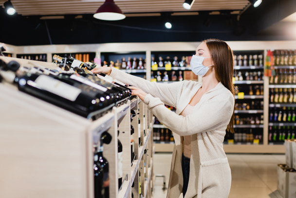 Mujer en máscara médica tomando botella de vino en primer plano borrosa en el supermercado  - Foto, Imagen