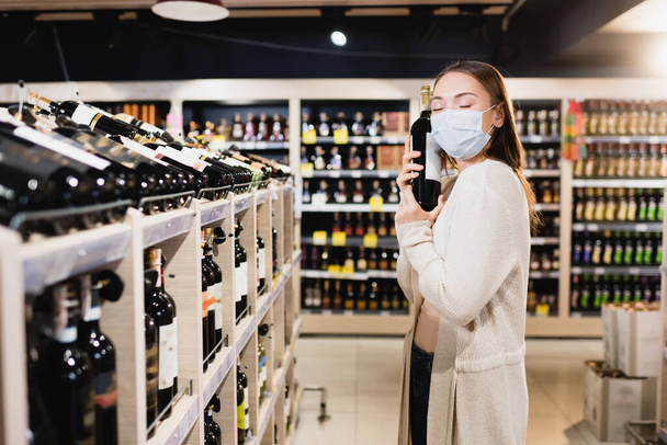 Woman in medical mask holding wine bottle in supermarket  - Fotoğraf, Görsel