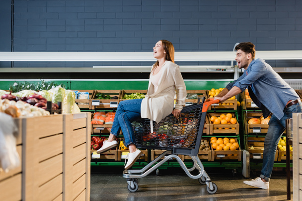 Coppia allegra rotolando sul carrello della spesa vicino alla spesa nel supermercato  - Foto, immagini