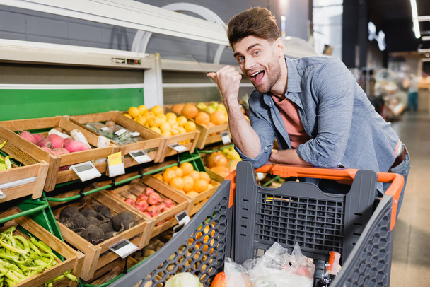 Веселый человек указывает на еду на прилавке возле корзины в супермаркете  - Фото, изображение