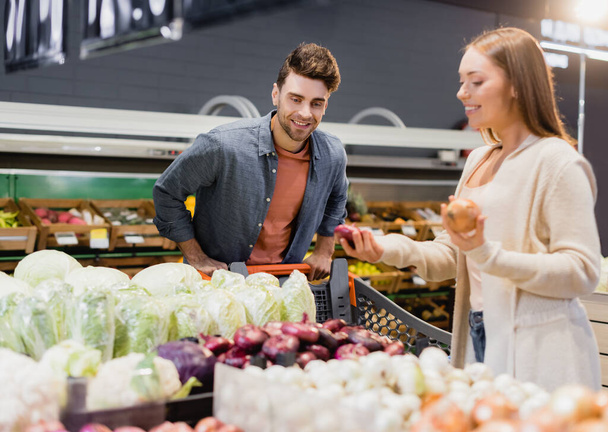 Sonriente hombre de pie cerca del carrito de la compra y novia con cebollas en primer plano borrosa en el supermercado  - Foto, Imagen