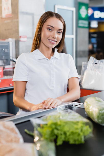 Lächelnde Kassiererin blickt in die Kamera in der Nähe von Lebensmitteln an der Supermarktkasse im verschwommenen Vordergrund  - Foto, Bild