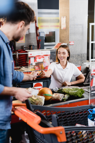 Smiling cashier taking credit card from customer near food on supermarket checkout - Zdjęcie, obraz