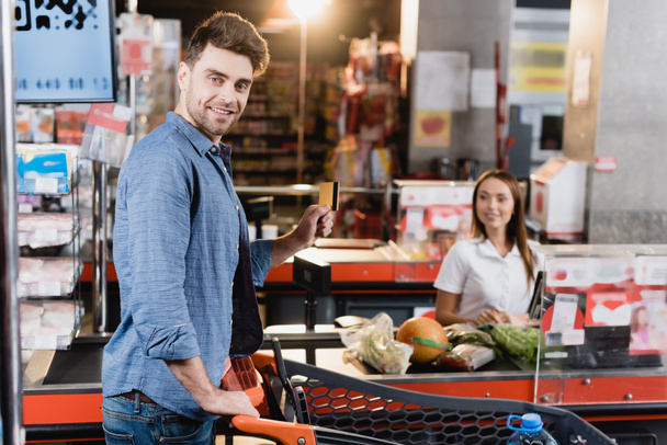 Smiling man holding credit card near shopping cart and supermarket checkout on blurred background  - 写真・画像