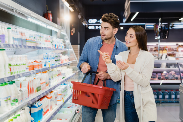 Frau mit Smartphone zeigt mit Finger auf Lebensmittel neben Freund mit Einkaufskorb im Supermarkt  - Foto, Bild