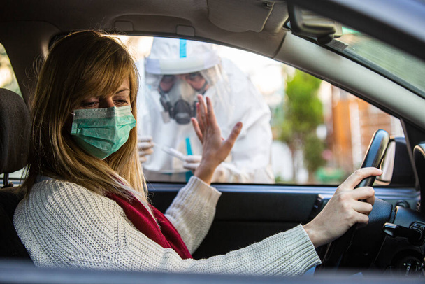Woman refuses medical worker trying to perform drive-thru COVID-19 test, taking nasal swab sample from patient through car window, PCR diagnostic, doctor in PPE holding test kit. She is holding passports. - Photo, Image