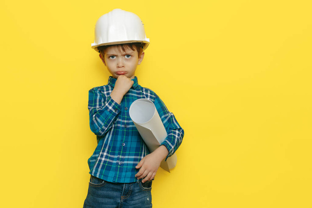 Serious little kid boy engineer or architect in a protective helmet holding construction plan over yellow background. - Foto, Imagem
