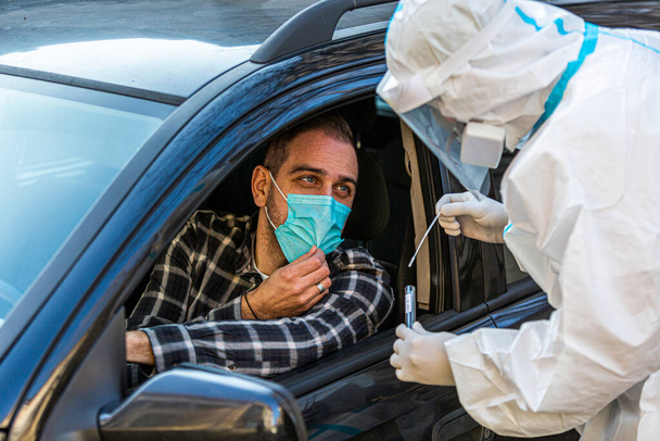 Мужчина, сидящий в машине и ожидающий, пока медработник проведет тест COVID-19, возьмет образец носового мазка через окно машины, ПЦР-диагностику коронавируса, врача в СИЗ с тестовым набором. - Фото, изображение