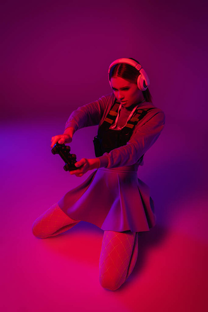KYIV, UKRAINE - NOVEMBER 27, 2020: young woman in wireless headphones holding joystick and playing video game on purple background  - Fotografie, Obrázek