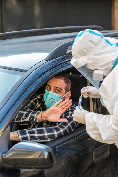 L'uomo rifiuta il medico che cerca di eseguire drive-thru COVID-19 test, prendendo campione di tampone nasale dal paziente attraverso il finestrino dell'auto, diagnostica PCR, medico in kit di prova PPE holding. Ha i passaporti.. - Foto, immagini