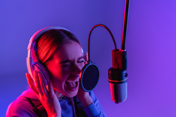 Frau mit drahtlosem Kopfhörer nimmt Lied auf, während sie in Mikrofon auf lila singt  - Foto, Bild