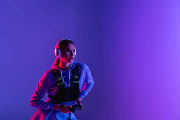 KYIV, UKRAINE - NOVEMBER 27, 2020: woman in wireless headphones holding gamepad and playing video game on purple  - Foto, immagini