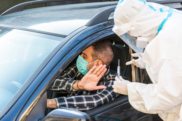 Mężczyzna odmawia pracownikowi medycznemu próby wykonania testu drive-thru COVID-19, pobrania próbki wymazu z nosa od pacjenta przez okno samochodu, diagnostyki PCR, lekarza w PPE posiadającego zestaw testowy. Trzyma paszporty.. - Zdjęcie, obraz