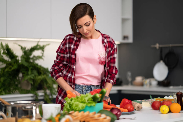 Cutting fresh vegetables pretty housewife cooking dinner wearing a plaid shirt. Cooking with passion young woman with short hair standing at modern kitchen. Healthy food leaving - concept - Foto, Imagem