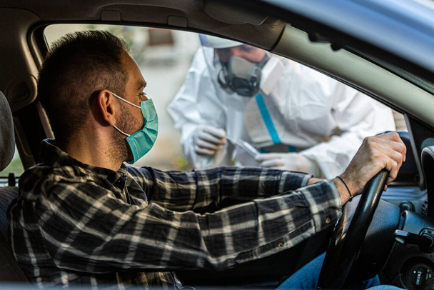 Мужчина, сидящий в машине и ожидающий, пока медработник проведет тест COVID-19, возьмет образец носового мазка через окно машины, ПЦР-диагностику коронавируса, врача в СИЗ с тестовым набором. - Фото, изображение
