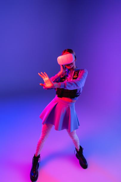 full length of young woman in virtual reality headset gesturing on purple background  - Foto, afbeelding