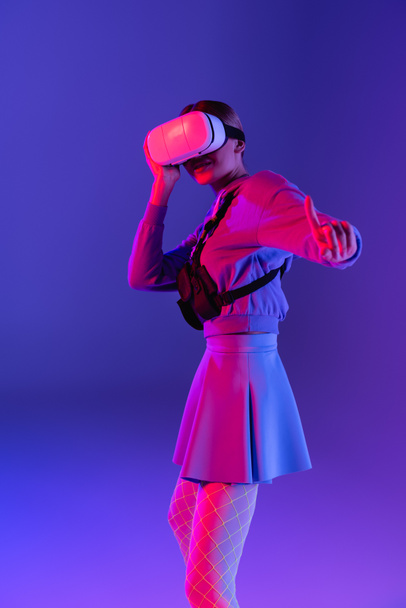 young woman in virtual reality headset pointing with finger on purple background  - Foto, Imagen