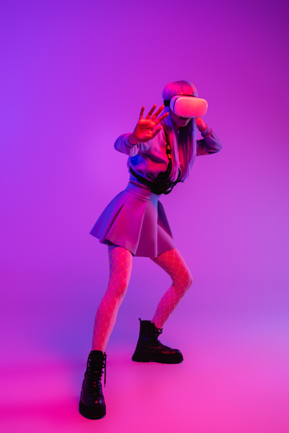 full length of young woman in virtual reality headset standing with outstretched hand on purple background  - Foto, imagen