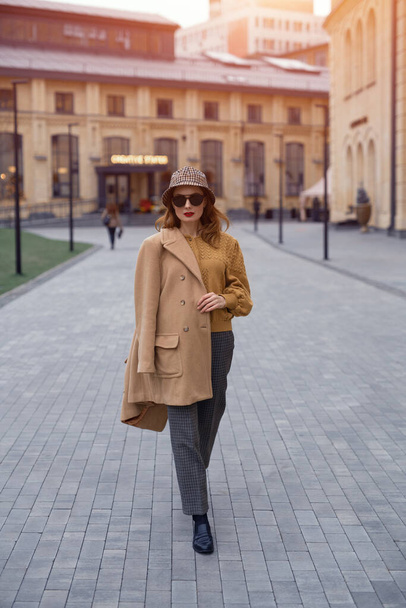 Confident fashion model young woman in sunglasses and hanging on a shoulder autumn beige coat walking on the street happily posing for the camera. Tinted photo - Фото, изображение