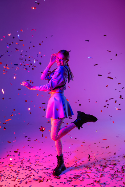 full length of happy woman in stylish outfit winking eye near falling confetti on purple - Photo, Image
