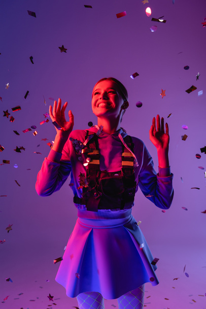 positive woman in stylish outfit near falling confetti on purple - Photo, Image