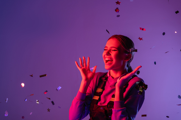 cheerful woman in stylish outfit near falling confetti on purple - Photo, Image
