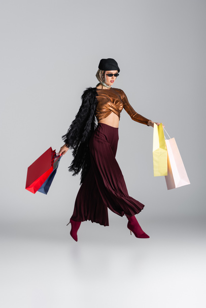 teljes hossza trendi fiatal nő napszemüveg és svájcisapka gazdaság bevásárló táskák, miközben lebegő szürke  - Fotó, kép