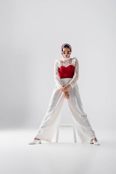 full length of stylish young woman in headscarf and sunglasses sitting on stool and posing on white  - Foto, imagen