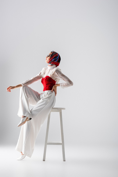 full length of stylish young woman in headscarf and sunglasses sitting with hand on hip on stool and looking away on white  - Fotografie, Obrázek