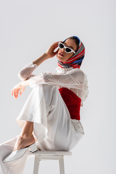 young woman in headscarf and stylish sunglasses sitting on stool while posing isolated on white  - Фото, зображення