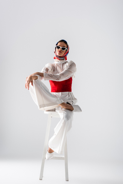 full length of stylish woman in headscarf and sunglasses sitting on stool while posing on white  - Foto, imagen