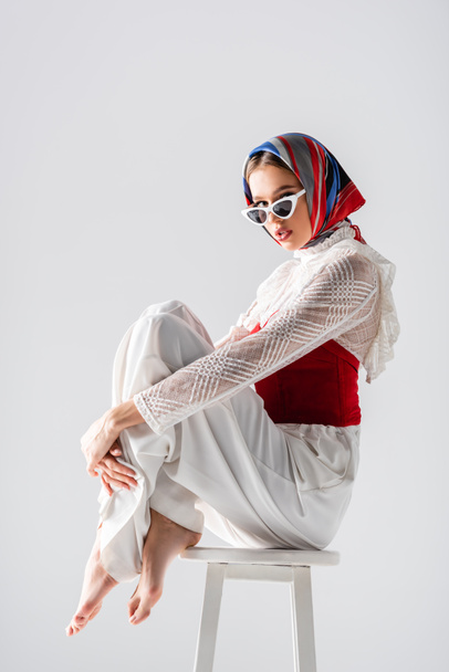 stylish woman in headscarf and sunglasses sitting on stool while posing isolated on white  - Fotoğraf, Görsel