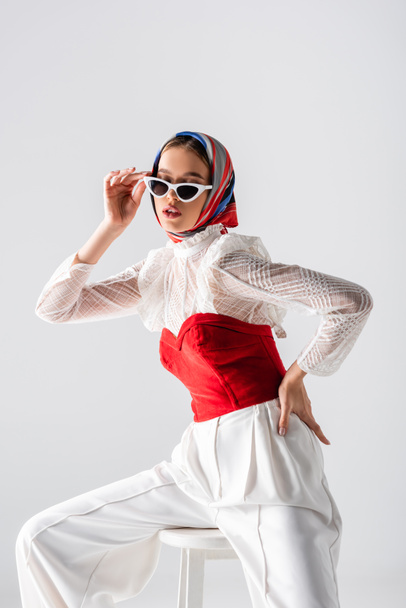 stylish woman in headscarf adjusting sunglasses and sitting on stool with hand on hip isolated on white  - Foto, immagini