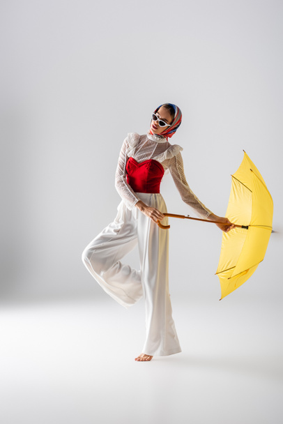 full length of stylish woman in headscarf and sunglasses holding yellow umbrella while posing on white  - Foto, immagini