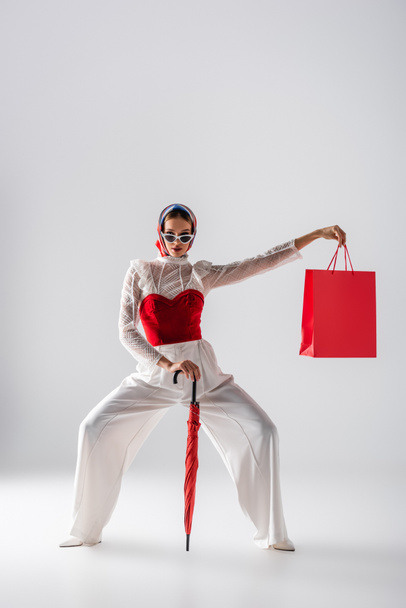 pełna długość kobiety w chustce i modne okulary przeciwsłoneczne z czerwonym parasolem i torbą na zakupy podczas pozowania na białym  - Zdjęcie, obraz