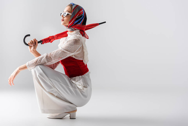 full length of young woman in headscarf and sunglasses holding red umbrella while sitting on white  - Фото, зображення