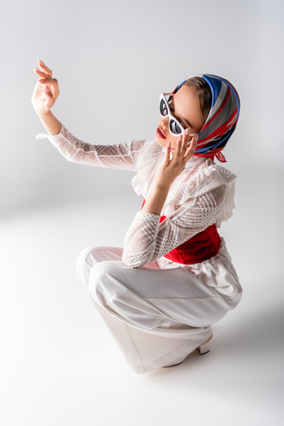 stylish woman in headscarf adjusting sunglasses, gesturing and sitting on white  - Foto, afbeelding