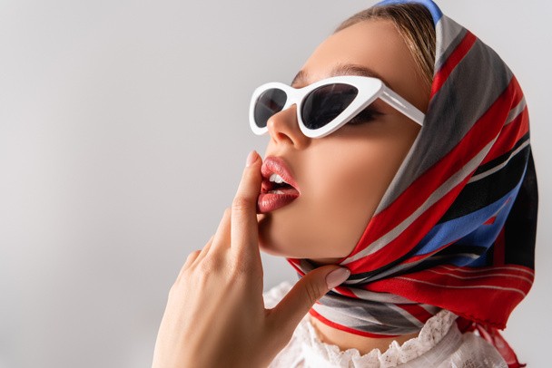 νέα και αισθησιακή γυναίκα με μαντίλα και γυαλιά ηλίου που αγγίζουν τα χείλη απομονωμένα σε λευκό - Φωτογραφία, εικόνα